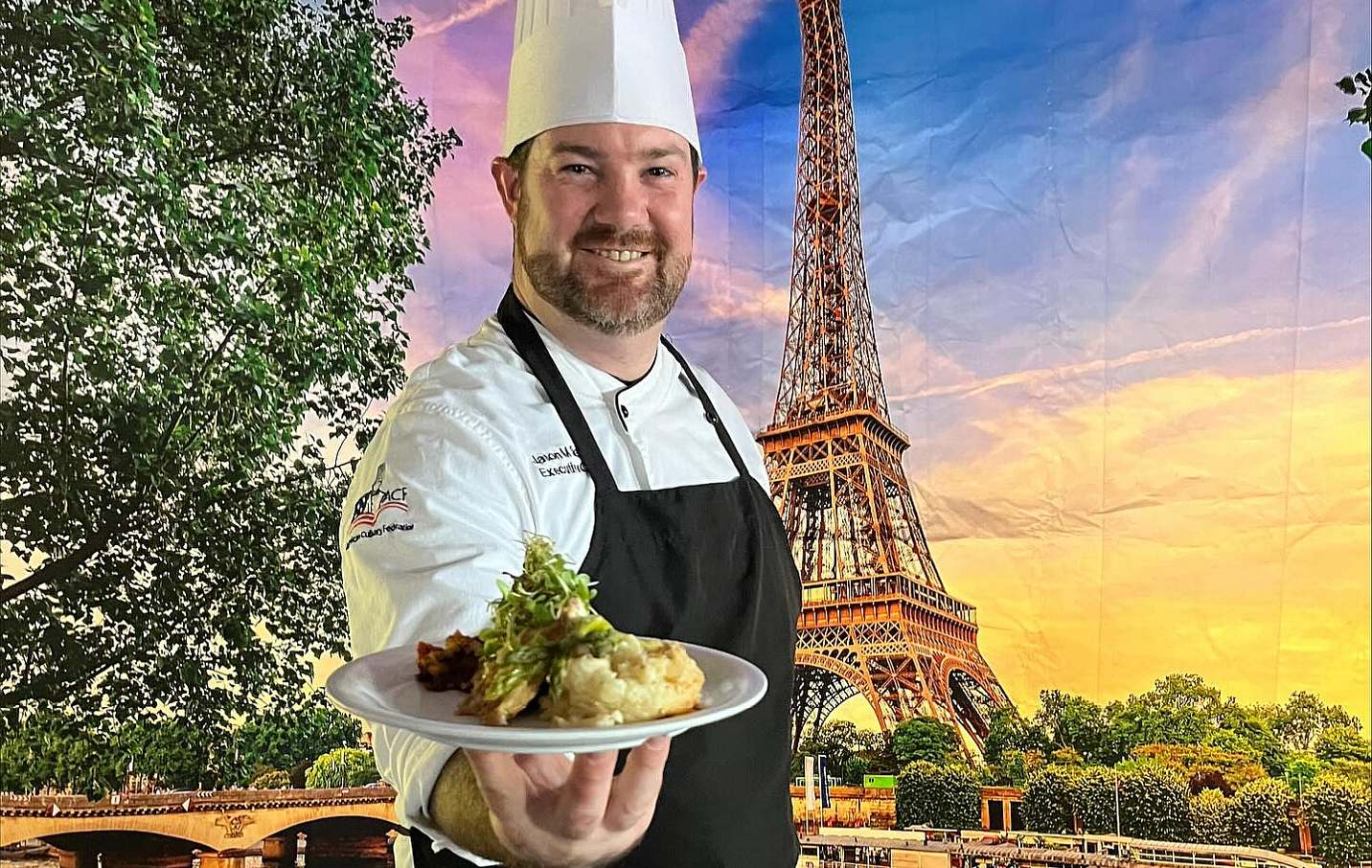 chef holding a plate of food in front of a mural of the Eiffel Tower